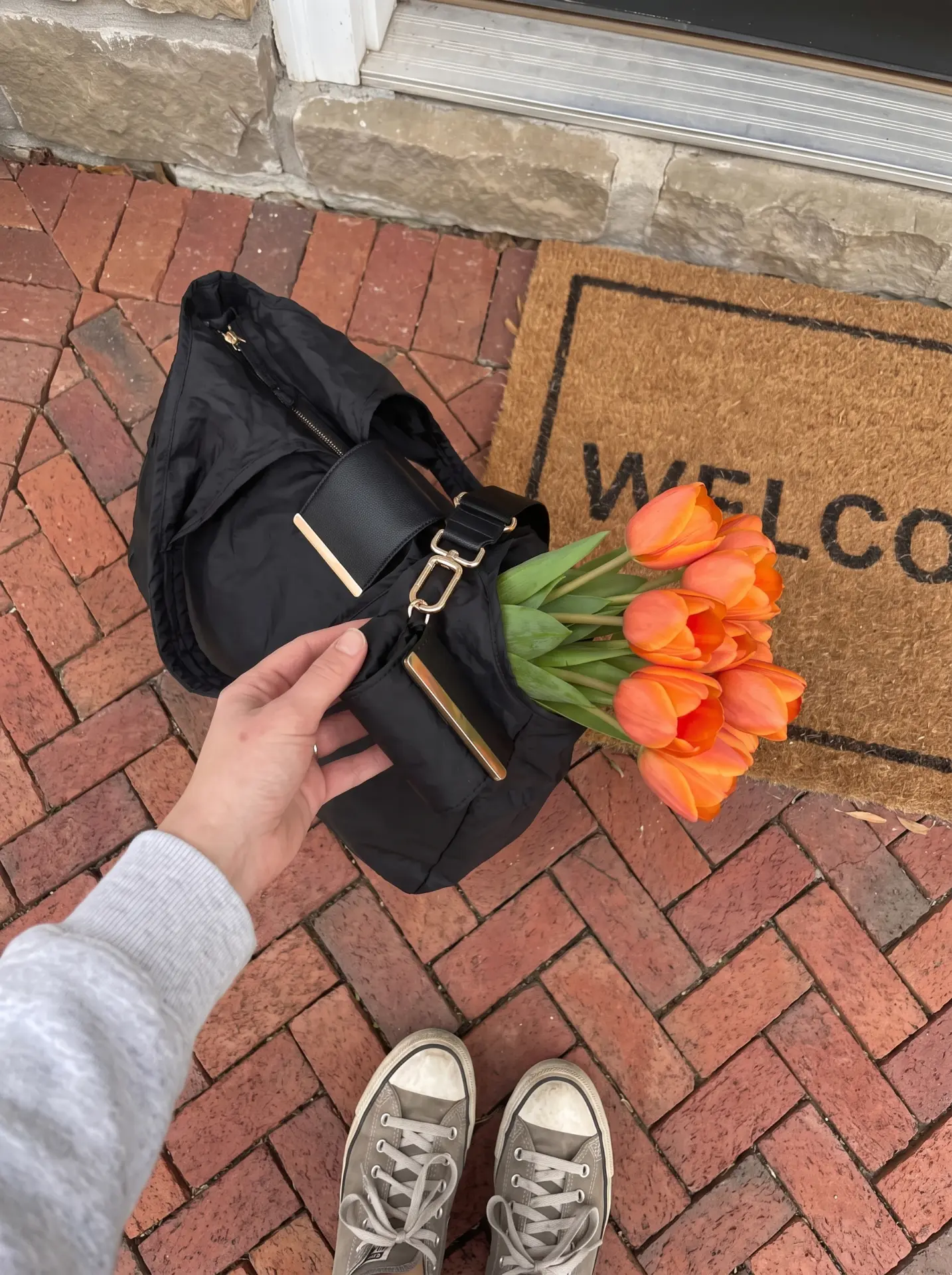 Hand holding the folded black Cliqué Tote pouch beside tulips on a doorstep
